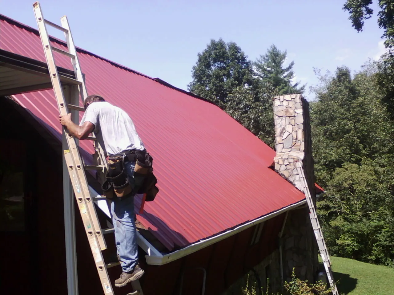 Roofer on a ladder beside a red metal roof and stone chimney — Soffit & Fascia Repair by Roofing Brothers in Queen